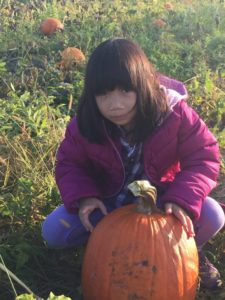 Paige kneeling on the ground behind a medium sized orange pumpkin. In the background is pumpkins