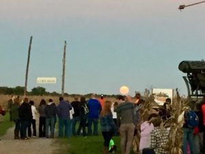 A crowd of people by the sign, corn maze, up in the sky there is a orange moonrise 