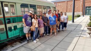Group photo in front of a Trolley Car that us offwhite at top, then an orange line, green strip, offwhite strip and another green strip. From left to right is Mary's family, Katie, Mary, Jane, Roland, Missy, Mark, Sam, Rick, Debbie, Roz 
